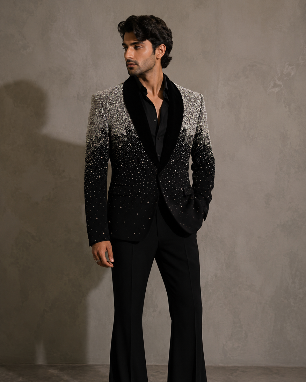 A man modeling a black and silver gradient sequined blazer with black satin lapels, paired with black flared trousers, against a textured gray studio background.